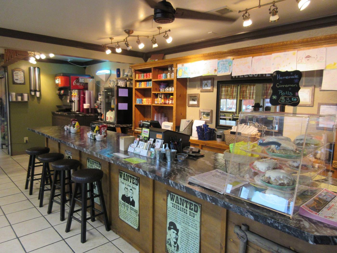 Inside the Hungry Hunter: a long counter with bar stools, a glass case of cinnamon rolls, wanted posters along the counter front and children's drawings pegged above.
