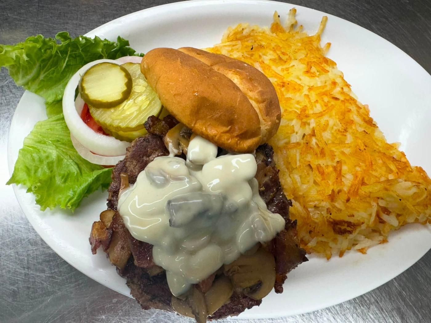A burger patty topped with mushrooms in swiss cheese sauce and bacon, with lettuce, tomato, onion, pickles and hash browns.