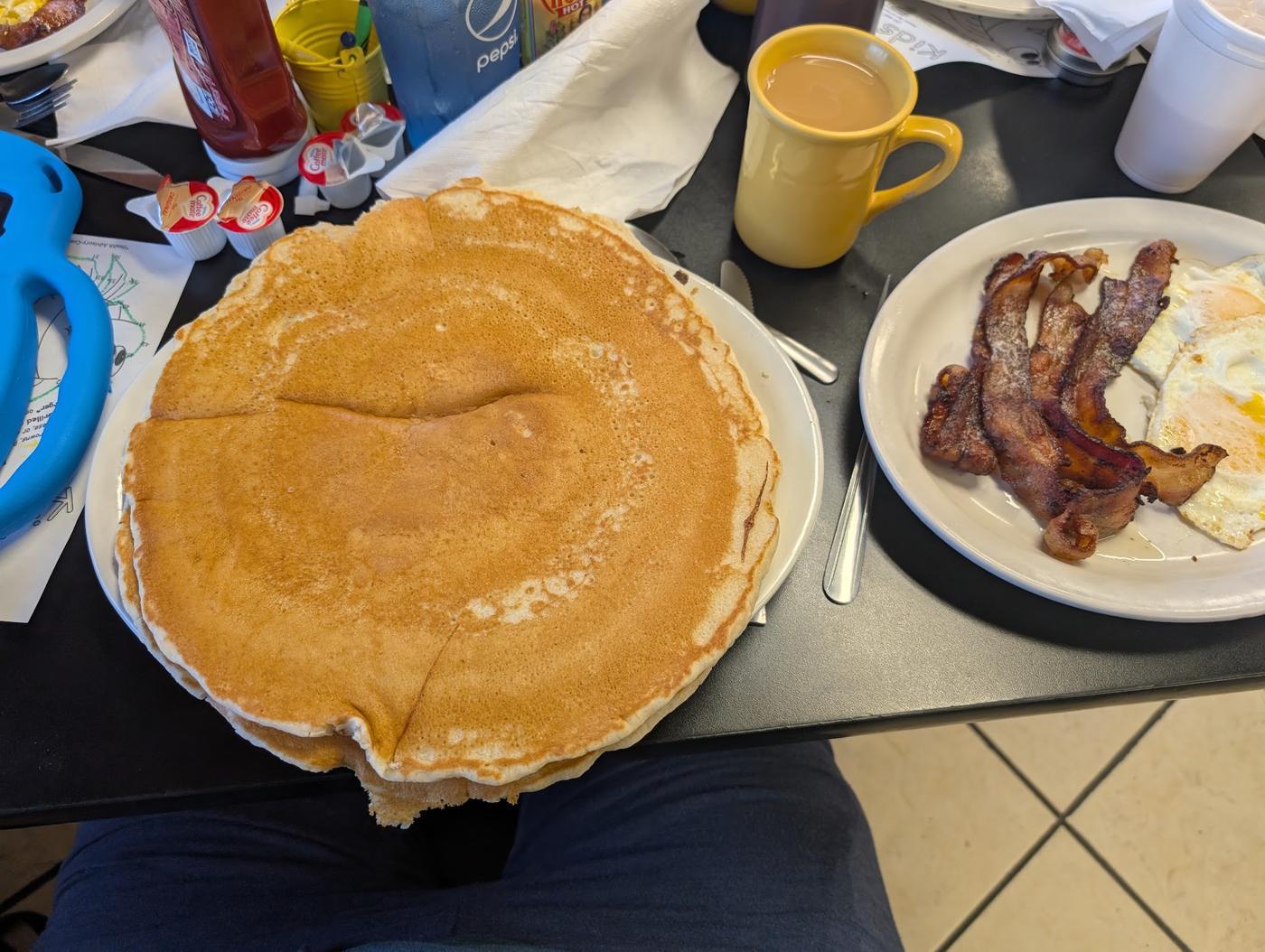 A single pancake so wide it hangs over every edge of the plate, beside a plate of bacon and eggs and a yellow mug of coffee.