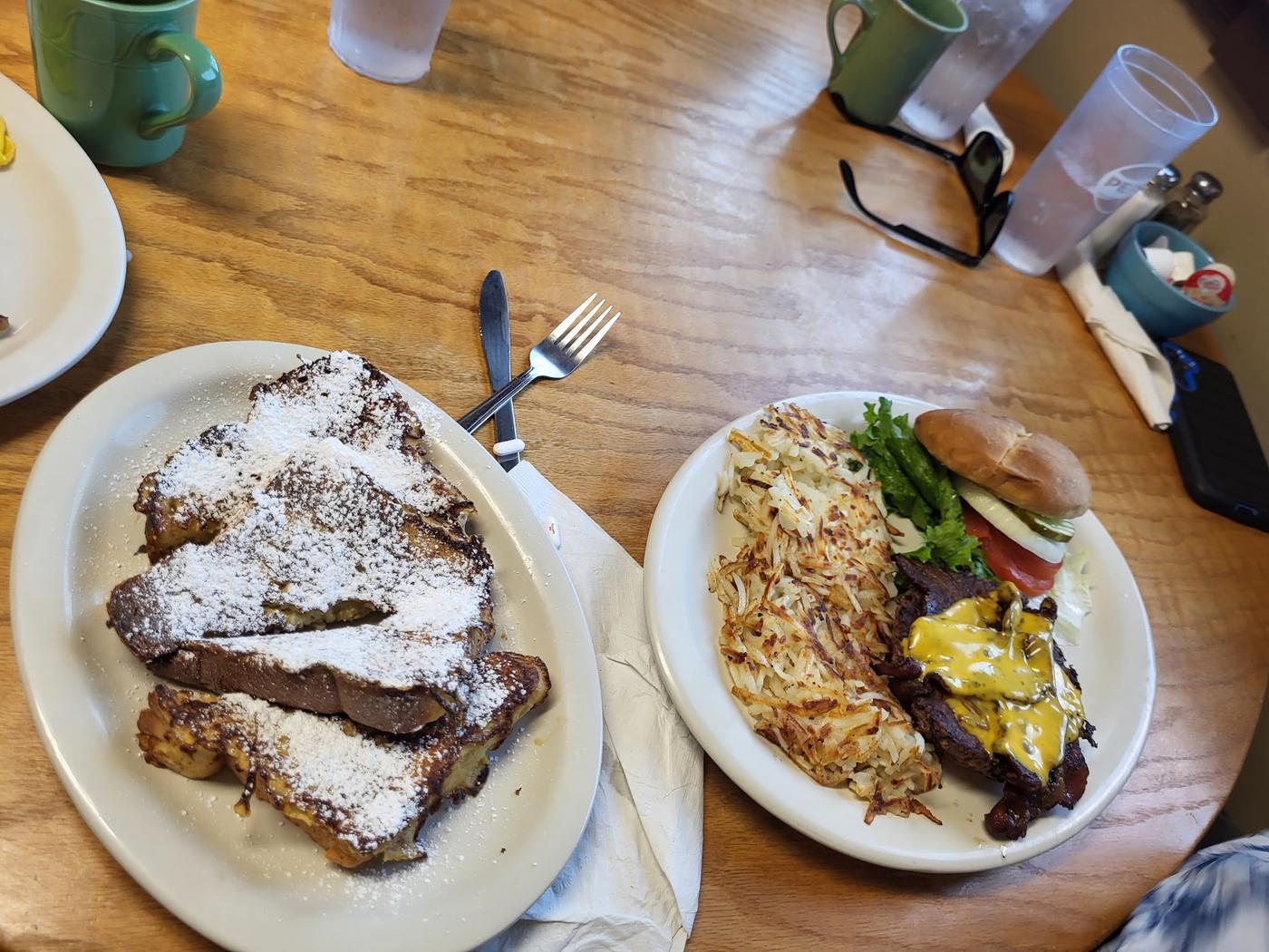 A wooden table with a platter of french toast under powdered sugar and a burger with hash browns, green mugs of coffee alongside.